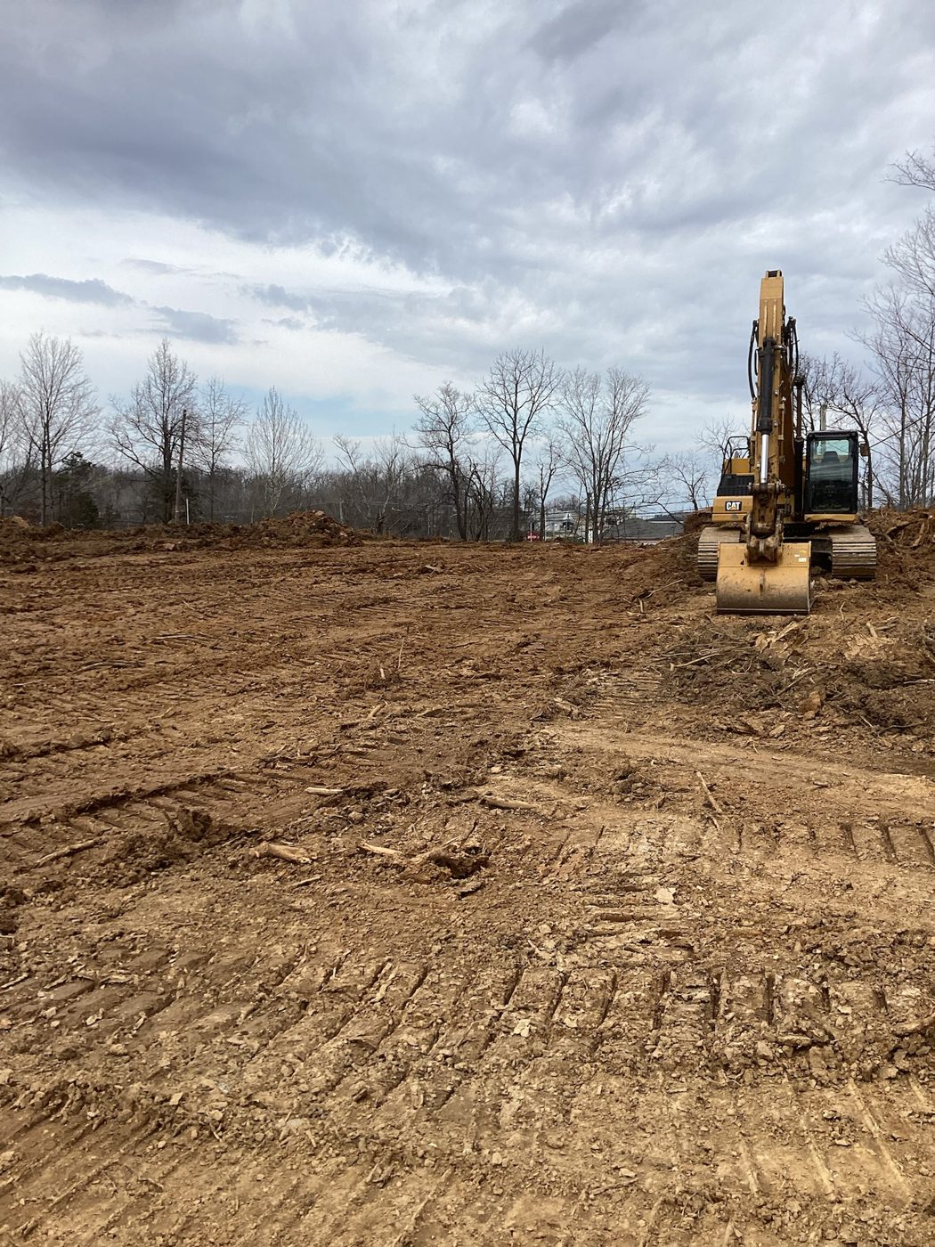 Excavator on the stripped lot after clearing, bare earth to the tree line