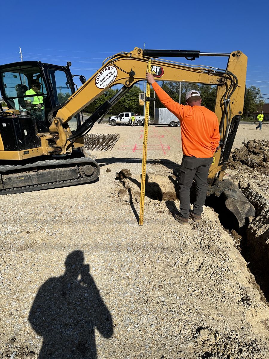 Excavator trenching for underground utilities while a crew member checks grade
